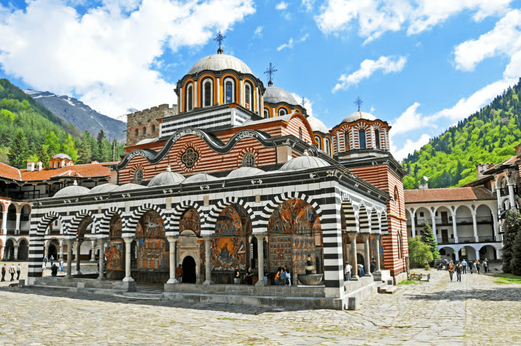 The main church of Rila Monastery with black and white striped arches, colourful frescoes and forested mountains behind