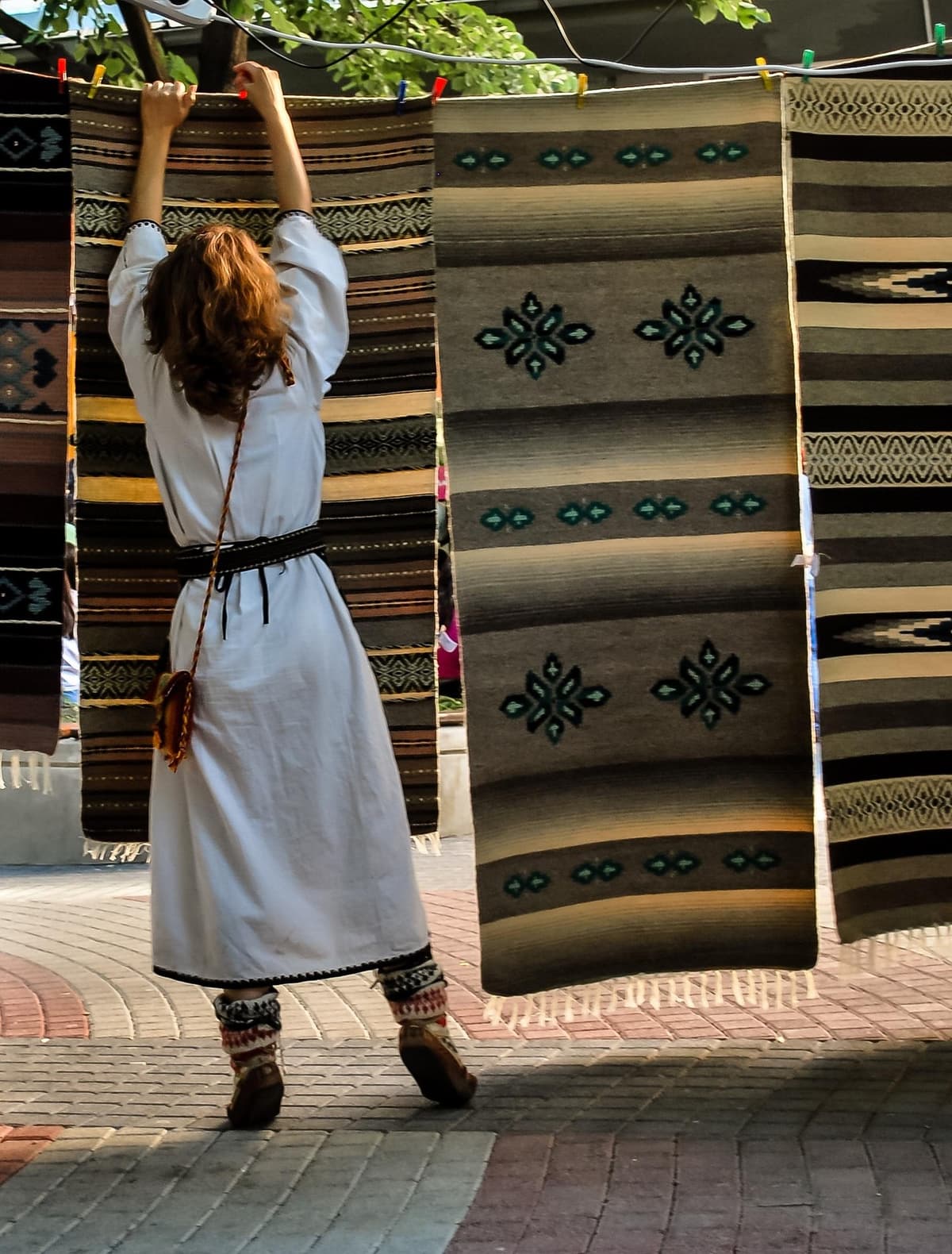 Woman in traditional white Bulgarian embroidered dress with black belt, displaying traditional Bulgarian kilim rugs with geometric patterns