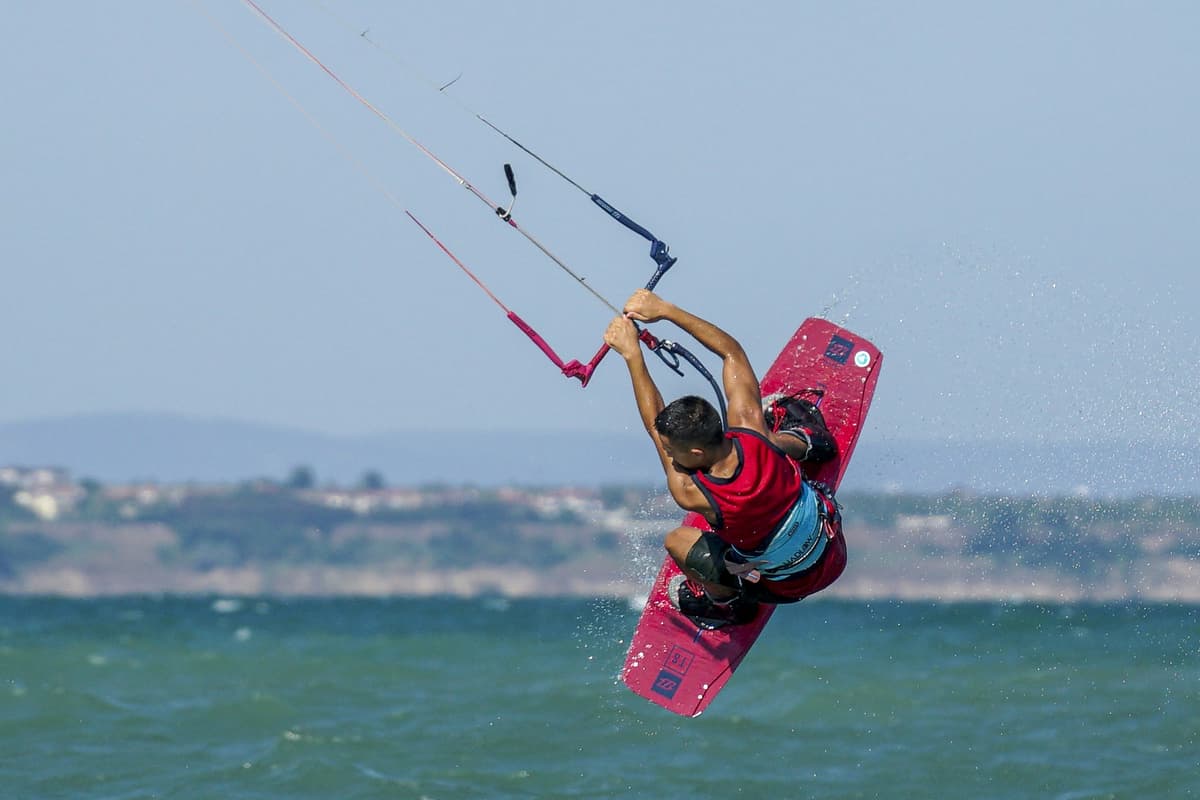 Kitesurfer in action above Bulgaria's Black Sea waters, leaping with red board against distant coastal buildings backdrop